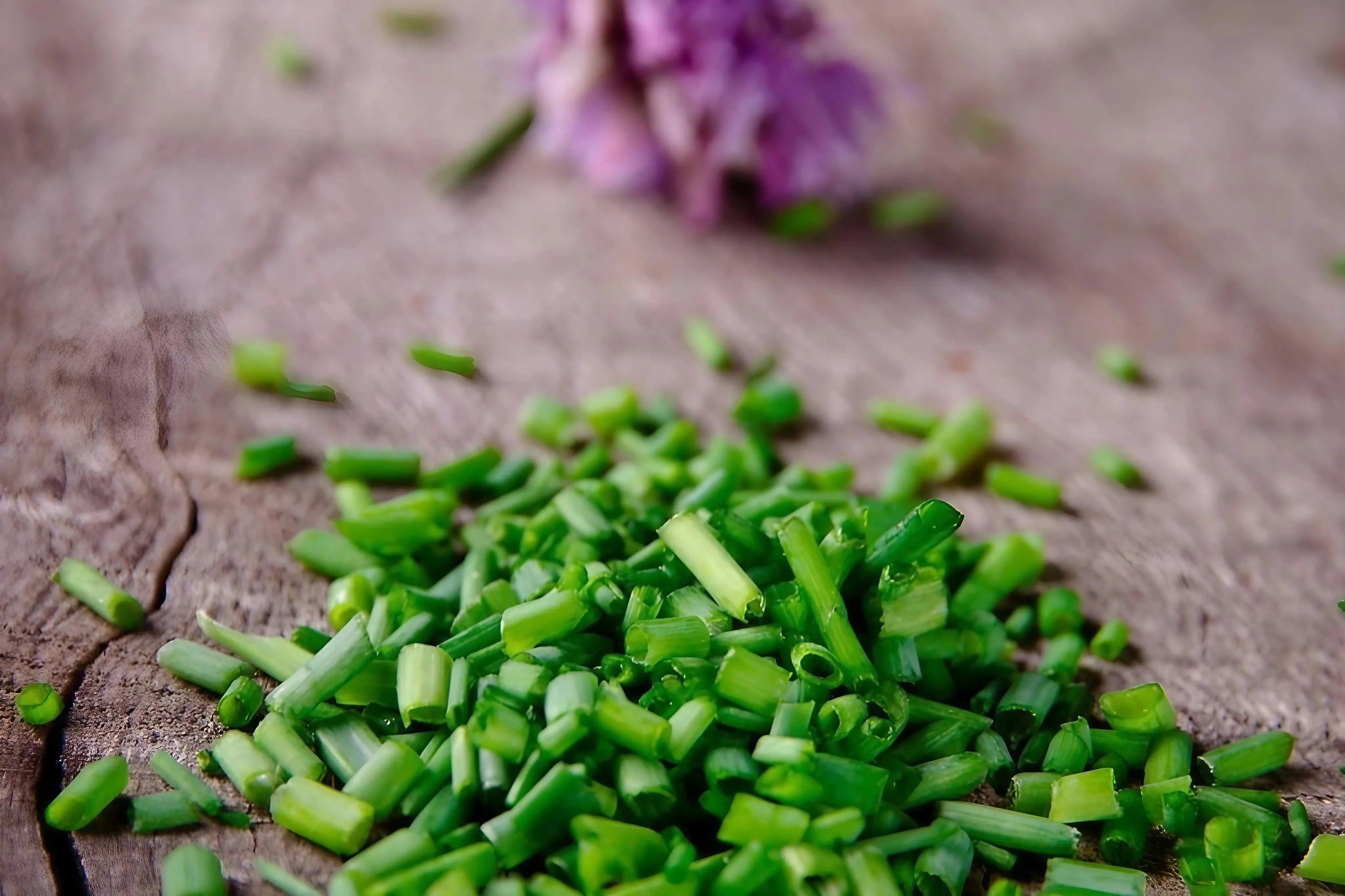 Chopped green chives on a wooden surface with purple flowers in the background