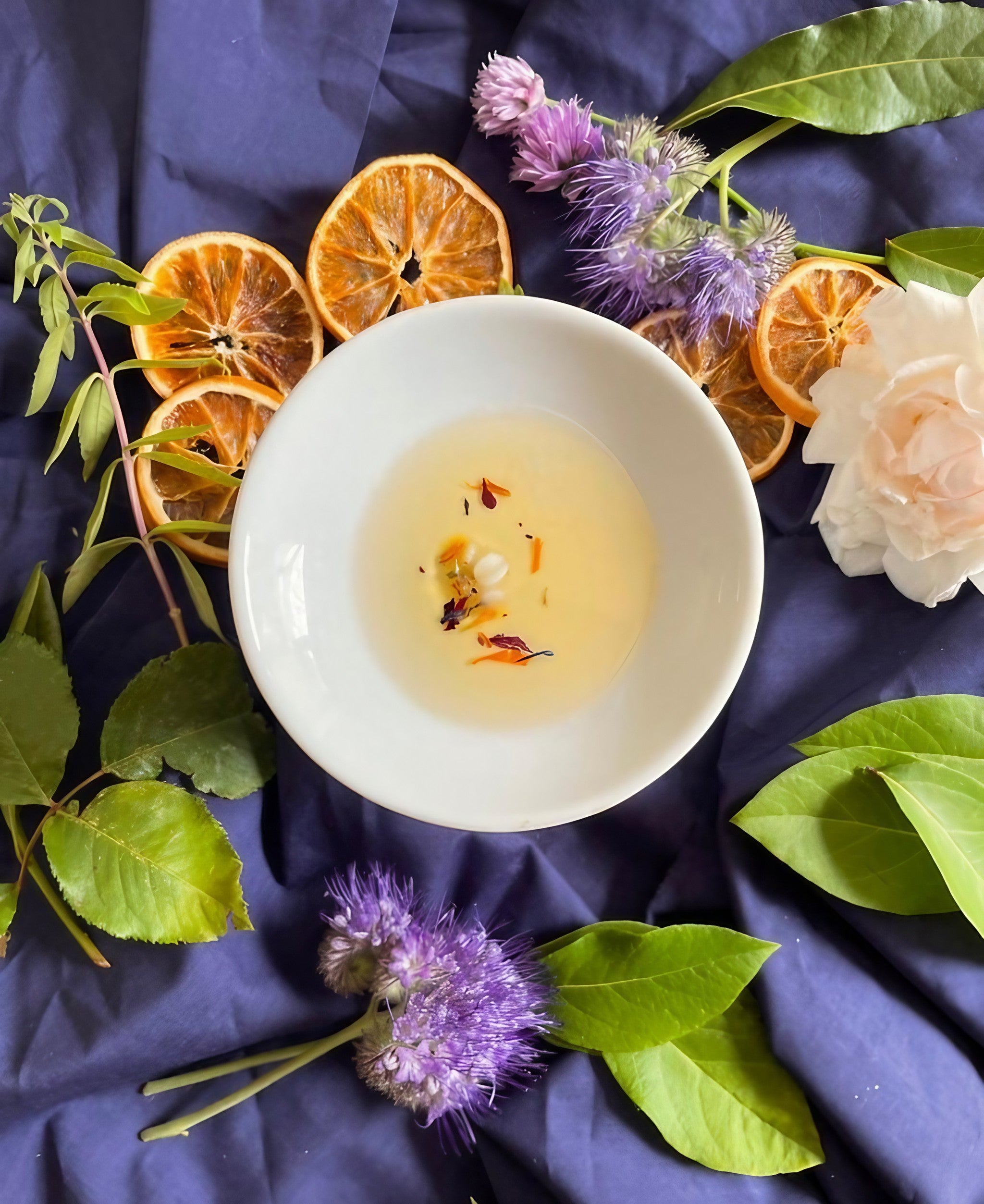 Bowl of scented oil surrounded by dried orange, flowers, and green leaves on a navy blue background.
