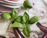Several fresh Basil Thai Siam Queen leaves from Bishy Barnabees Cottage Garden Ltd are arranged on a surface with a blurred maroon and white background, creating a striking contrast.