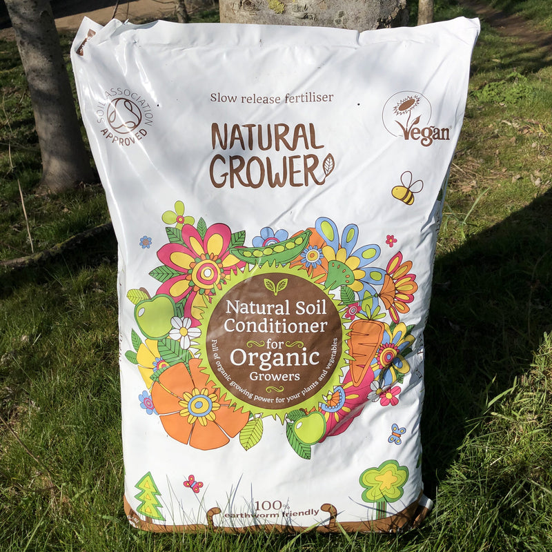 A 50L bag of Natural Grower Natural Plant Feed stands on grass, featuring colorful flower illustrations and labels showing it’s a slow-release, vegan organic soil conditioner approved by the Soil Association.