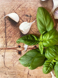 Bishy Barnabees Cottage Garden Ltd’s Basil Classic Italian brings vibrant color to the background, with fresh green leaves alongside two garlic cloves on a rustic wooden cutting board.