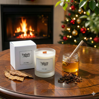 A Tadpole & Piglet Dark Honey & Tobacco Scented Candle sits on a wooden table with its box, honey jar, dried leaves, and loose tea. A lit fireplace and decorated Christmas tree glow in the cozy background.