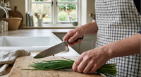 Wearing a checkered apron, a person slices fresh Chives from Bishy Barnabees Cottage Garden Ltd on a wooden cutting board in a bright kitchen with potted herbs, a window, and a bowl of eggs nearby.