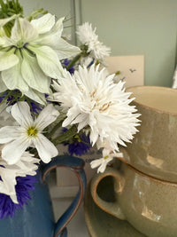 Cornflower Snowman by Bishy Barnabees Cottage Garden features white and purple double blooms in a blue vase, displayed beside stacked cream ceramic cups and saucers against a light background.
