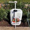 A large white 10L Jerry Can Dispensing Pump by Natural Grower, labeled Organic Fertiliser, stands on soil among green plants and flowers in a garden, ready to provide liquid feed.