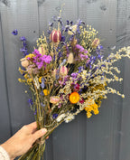 A hand displays a Bishy Barnabees Cottage Garden Ltd Dried Flower Bouquet with purple, yellow, pink, and white blooms against a dark gray wooden background.