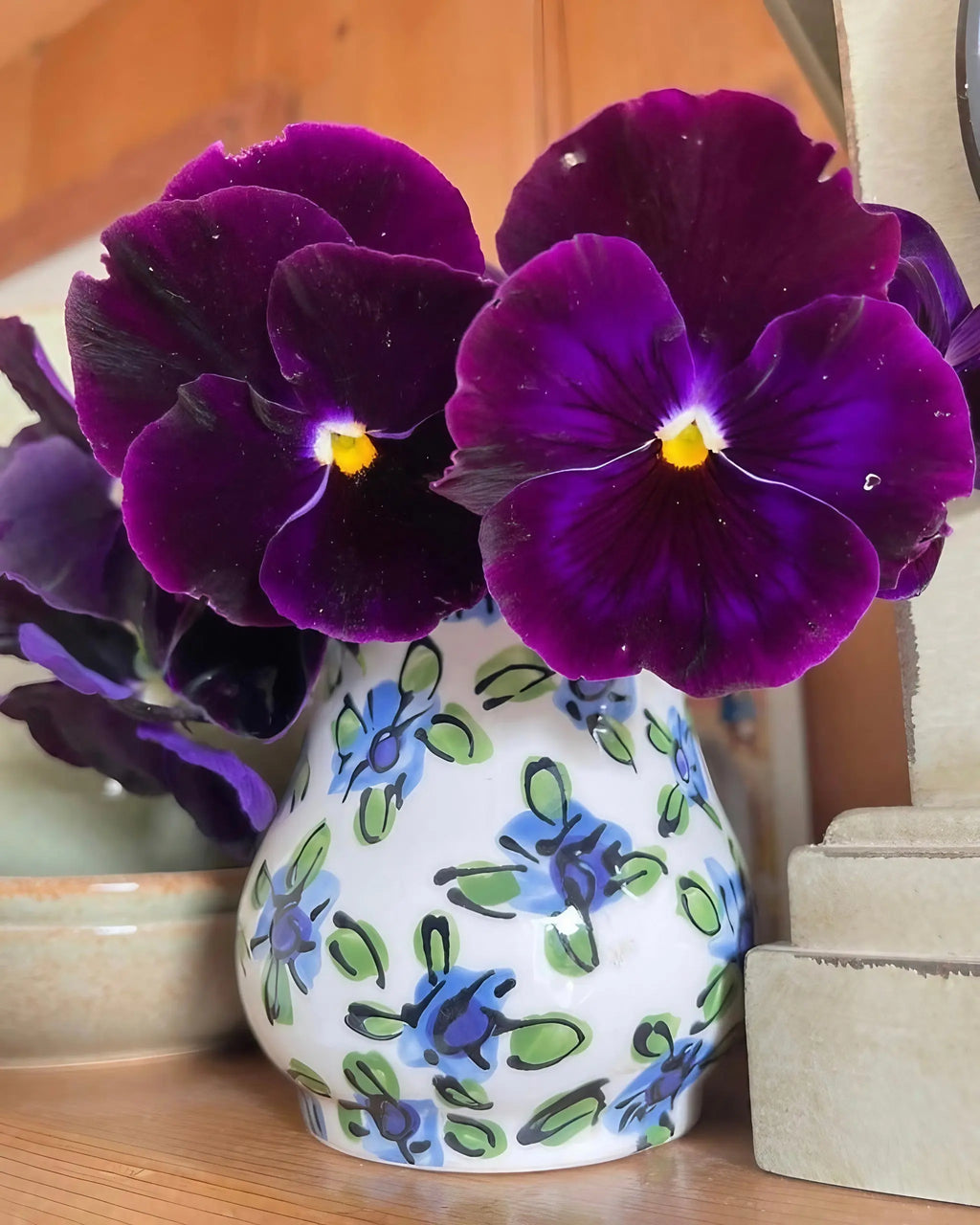 A small white ceramic vase decorated with blue and green flowers holds deep purple pansies from Bishy Barnabees Cottage Garden’s Patio Flower Garden Collection, set on a wooden surface beside a beige stone object.