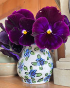 A small white ceramic vase decorated with blue and green flowers holds deep purple pansies from Bishy Barnabees Cottage Garden’s Patio Flower Garden Collection, set on a wooden surface beside a beige stone object.