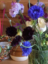 A vase of vibrant blue, purple, and white Cornflower Black Ball blooms from Bishy Barnabees Cottage Garden Ltd sits on a shelf with a pot, teacup, and beaded heart—perfect for adding cottage garden charm to your home.