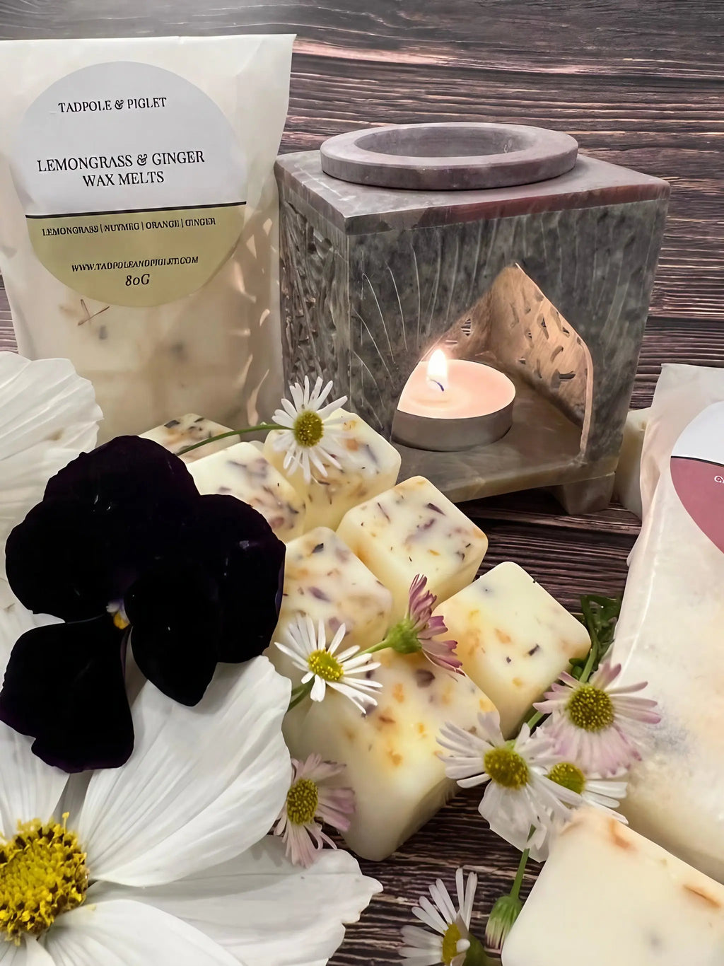 The Moroccan Arch Soapstone Wax Melt Burner by Bishy Barnabees Cottage Garden glows with a tealight behind lemongrass and ginger wax melts. White and purple flowers surround a labeled wax melts bag on the left, set against a cozy wooden background.