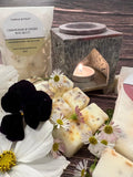 The Moroccan Arch Soapstone Wax Melt Burner by Bishy Barnabees Cottage Garden glows with a tealight behind lemongrass and ginger wax melts. White and purple flowers surround a labeled wax melts bag on the left, set against a cozy wooden background.