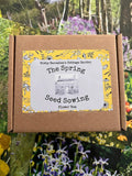 A brown cardboard box labeled "The Spring Seed Sowing Flower Box" by Bishy Barnabees Cottage Garden, with a yellow floral border, sits on flower photos—a perfect kit to start your cut flower garden dreams.