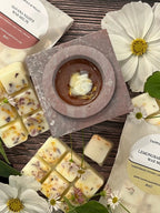 A wax-filled bowl sits atop the Bishy Barnabees Cottage Garden Moroccan Arch Soapstone Wax Melt Burner, surrounded by floral wax melts, white flowers, and two “HAVANA NIGHTS” and “LEMONGRASS” labeled wax melt packages on a brown surface.