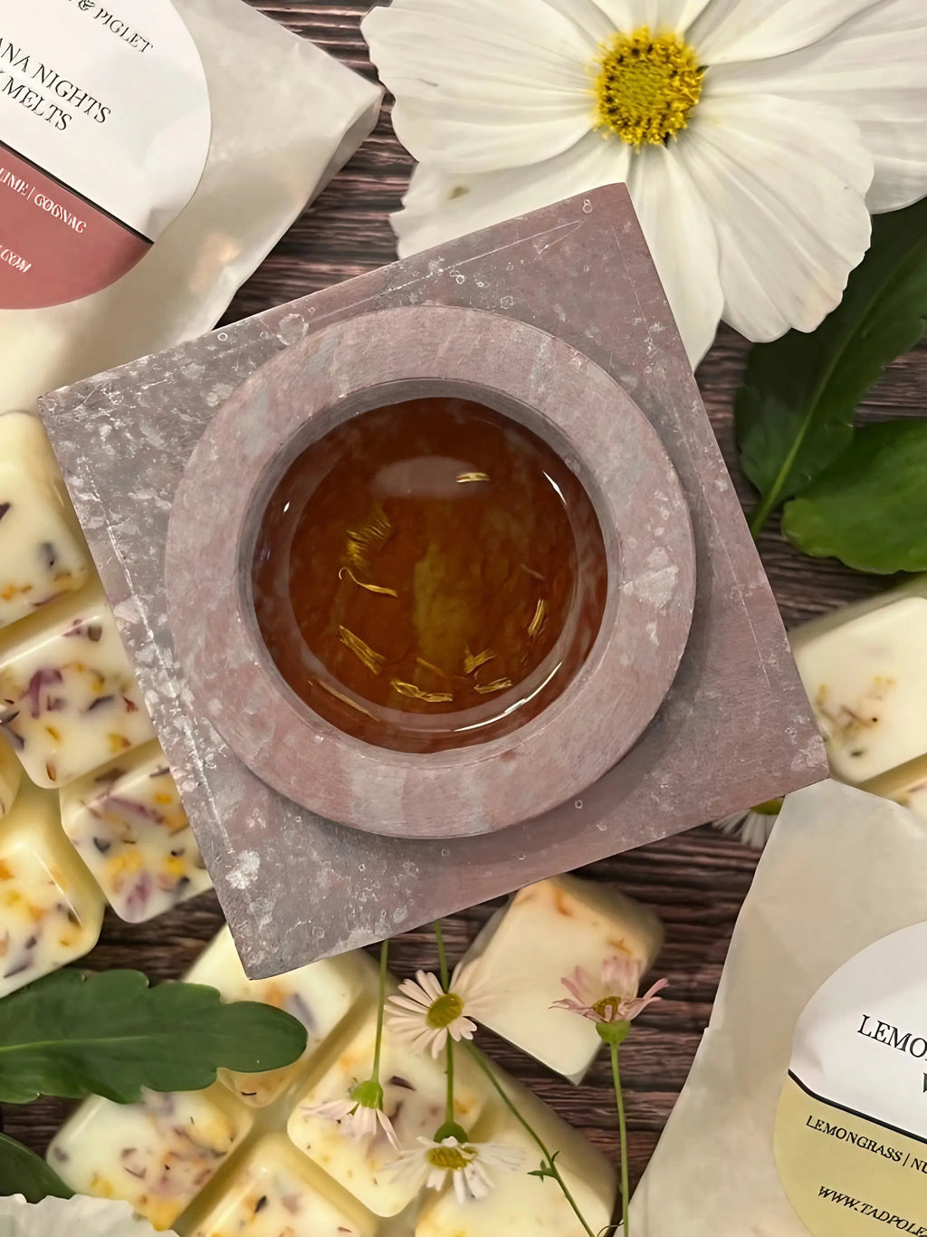 An amber liquid fills the Moroccan Arch Soapstone Wax Melt Burner by Bishy Barnabees Cottage Garden, surrounded by floral wax melts, green leaves, and white flowers on a wooden surface.