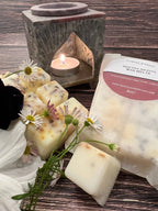 Soy wax cubes with dried herbs and flowers are arranged with daisies beside a “HAVANA NIGHTS WAX MELTS” package. A lit tealight adds elegance inside the Bishy Barnabees Cottage Garden Moroccan Arch Soapstone Wax Melt Burner.