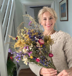 A smiling woman in a cream sweater holds a large Bishy Barnabees Cottage Garden Ltd Dried Flower Bouquet while standing indoors by a white staircase and framed art.