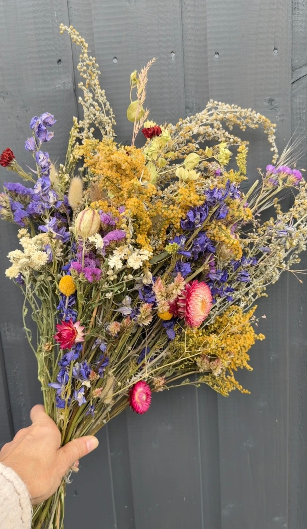 A hand holds a Bishy Barnabees Cottage Garden Ltd Dried Flower Bouquet featuring vibrant yellow, purple, red, and white blooms against a dark gray wooden background.