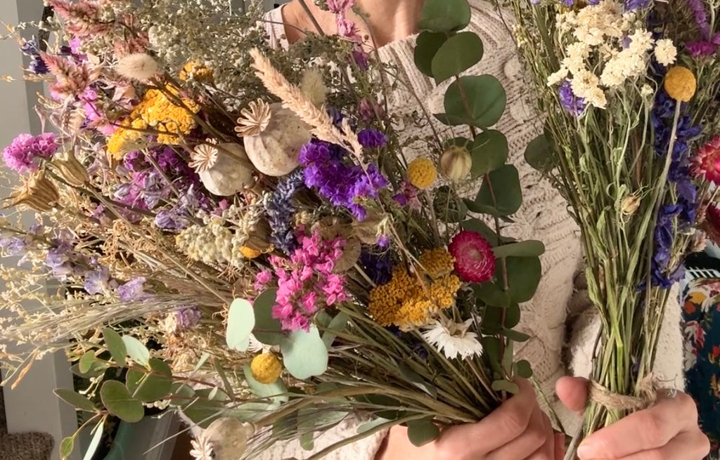 A person in a cream sweater holds two Dried Flower Bouquets by Bishy Barnabees Cottage Garden Ltd, showcasing sustainable yellow, purple, pink, and white blooms with green eucalyptus leaves and grasses.