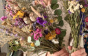 A person in a cream sweater holds two Dried Flower Bouquets by Bishy Barnabees Cottage Garden Ltd, showcasing sustainable yellow, purple, pink, and white blooms with green eucalyptus leaves and grasses.