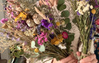 A person in a cream sweater holds two Dried Flower Bouquets by Bishy Barnabees Cottage Garden Ltd, showcasing sustainable yellow, purple, pink, and white blooms with green eucalyptus leaves and grasses.