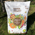 A 50L bag of Natural Grower Natural Plant Feed stands on grass, featuring colorful flower illustrations and labels showing it’s a slow-release, vegan organic soil conditioner approved by the Soil Association.