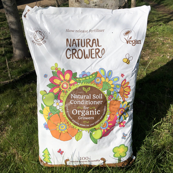 A 50L bag of Natural Grower Natural Plant Feed stands on grass, featuring colorful flower illustrations and labels showing it’s a slow-release, vegan organic soil conditioner approved by the Soil Association.