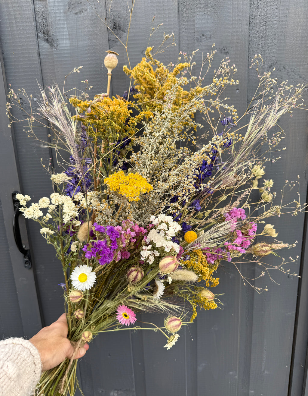 A hand displays Bishy Barnabees Cottage Garden Ltd's Dried Flower Bouquets, showcasing vibrant yellow, white, purple, and pink blooms in front of a gray wooden door.