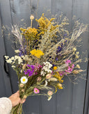 A hand displays Bishy Barnabees Cottage Garden Ltd's Dried Flower Bouquets, showcasing vibrant yellow, white, purple, and pink blooms in front of a gray wooden door.