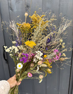 A hand displays Bishy Barnabees Cottage Garden Ltd's Dried Flower Bouquets, showcasing vibrant yellow, white, purple, and pink blooms in front of a gray wooden door.
