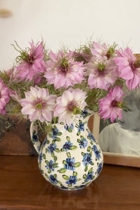 A ceramic jug from Bishy Barnabees Cottage Garden holds Nigella Mulberry Rose (Love-in-a-mist) blooms with pink flowers and burgundy-striped seed pods, sitting on a wooden surface beside a sepia-toned framed photo.