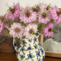 A ceramic jug from Bishy Barnabees Cottage Garden holds Nigella Mulberry Rose (Love-in-a-mist) blooms with pink flowers and burgundy-striped seed pods, sitting on a wooden surface beside a sepia-toned framed photo.