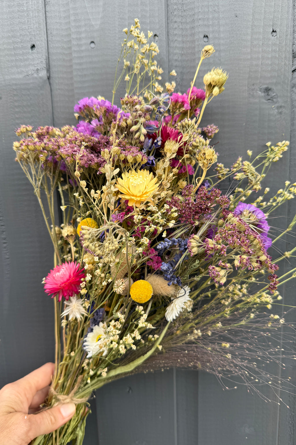 A hand holds a Bishy Barnabees Cottage Garden Ltd Dried Flower Bouquet with vibrant yellow, pink, purple, and white blooms, set against a gray wooden background.