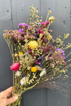 A hand holds a Bishy Barnabees Cottage Garden Ltd Dried Flower Bouquet with vibrant yellow, pink, purple, and white blooms, set against a gray wooden background.