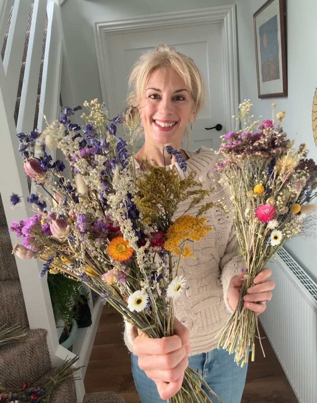 A smiling woman in a light sweater and blue jeans holds up two large Bishy Barnabees Cottage Garden Ltd Dried Flower Bouquets in purple, yellow, pink, and white tones while standing indoors.