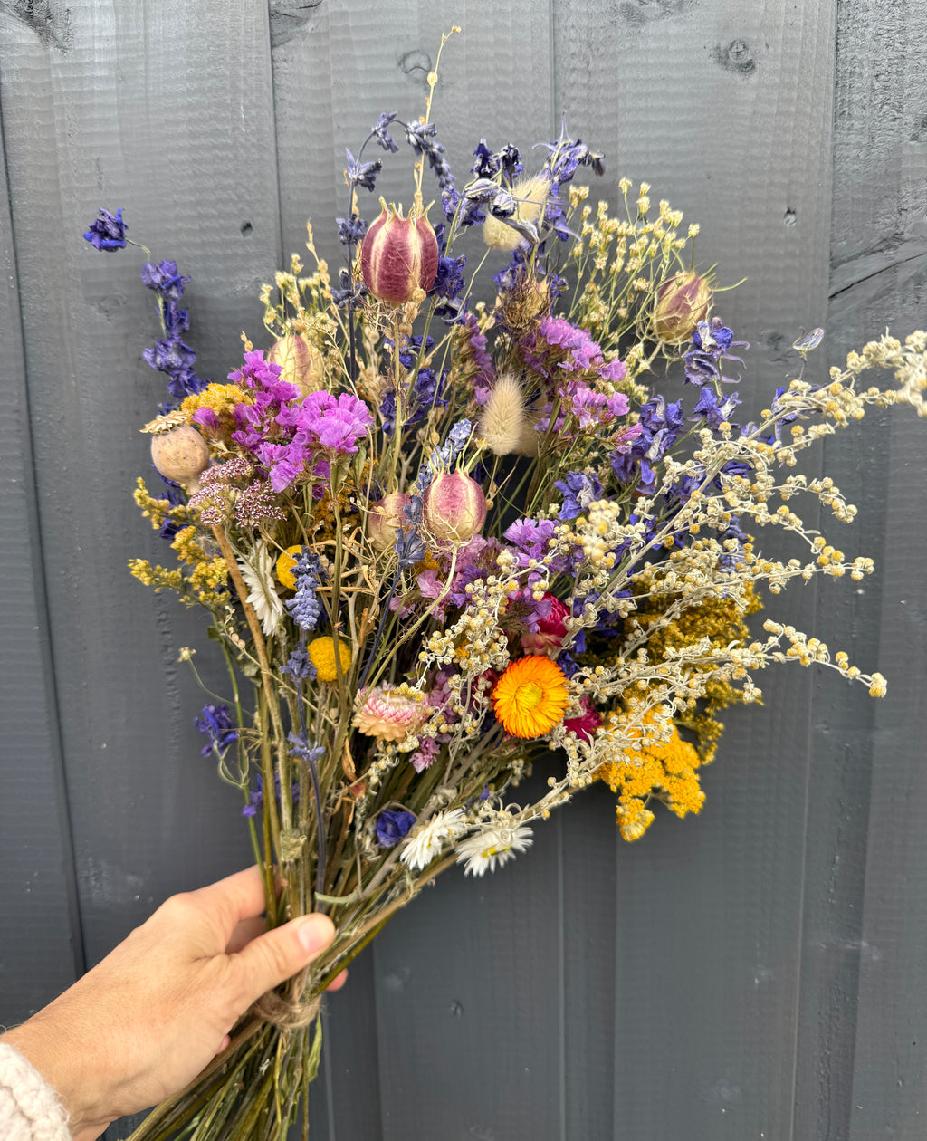 A hand holds a Bishy Barnabees Cottage Garden Ltd Dried Flower Bouquet in purple, yellow, pink, and white against a gray wooden background.