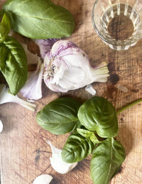 Bishy Barnabees Cottage Garden Ltd’s Basil Classic Italian, fresh basil leaves, and separated garlic cloves rest on a rustic wooden surface beside a clear glass, accented by scattered garlic peels and a softly blurred background.