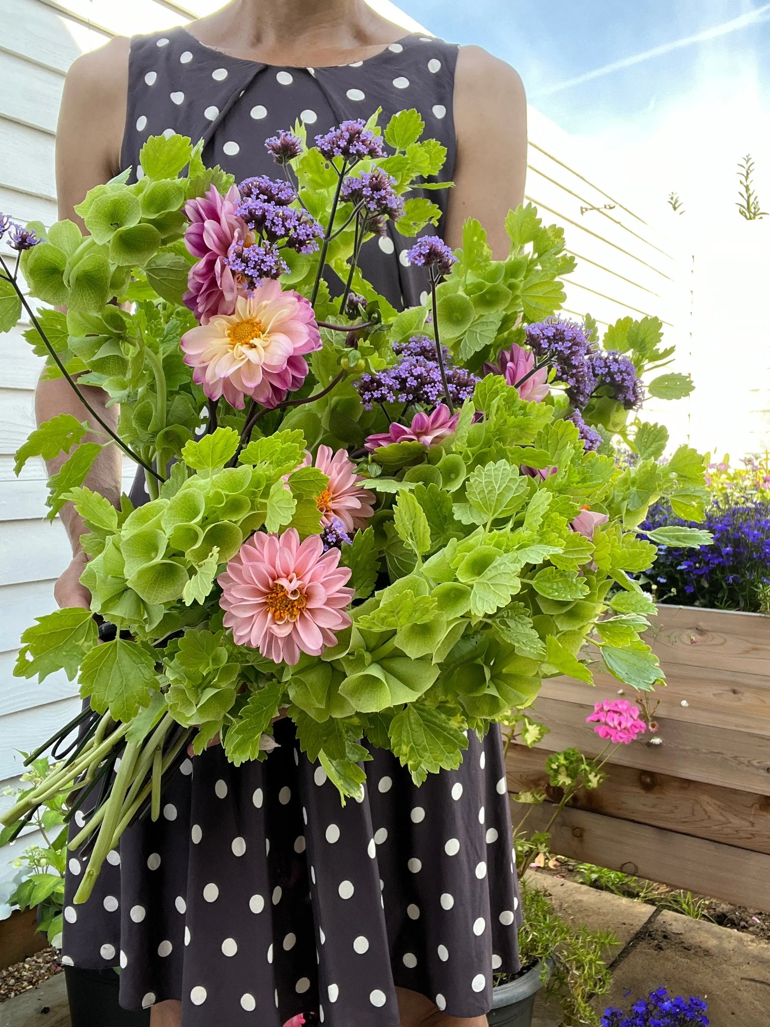 A person in a sleeveless polka dot dress holds a large bouquet featuring Bells of Ireland by Bishy Barnabees Cottage Garden Ltd, standing outdoors near white siding with a wooden planter box in the background.