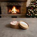 A round box of assorted chocolates sits open on a gray table beside Tadpole & Piglet Coffee Caramel Botanical Wax Melts, with a fireplace burning in the background and a lit Christmas tree to the right.