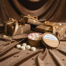 A round box of Tadpole & Piglet Coffee Caramel Botanical Wax Melts, some shaped like roses, sits open on brown velvet near wrapped gifts with gold ribbons and scattered gold star confetti.