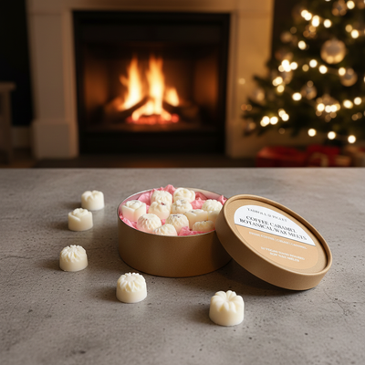 A round box of white chocolates sits open on the table, candies scattered nearby. Tadpole & Piglet Coffee Caramel Botanical Wax Melts fill the room with a warm aroma as a fireplace and Christmas tree create a cozy holiday mood.