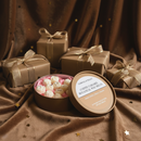 A round box of white truffles beside three brown gift boxes with gold ribbons sits on brown fabric with gold star confetti—perfectly paired with Tadpole & Piglet’s Coffee Caramel Botanical Wax Melts for an elegant touch.