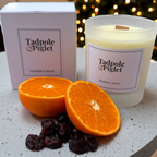 A Tadpole & Piglet Cranberry & Orange Scented Candle with a wooden wick sits beside its box, surrounded by orange halves and dried cranberries on a light surface, against a softly glowing backdrop.