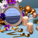 A round box of Tadpole & Piglet Damson & Rose Botanical Wax Melts rests on a blue surface with candles, gold spoons, flowers, gold ornaments, and ribbon. These vegan, hand-poured melts feature a fruity floral scent.