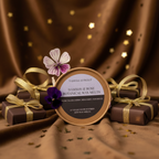 A round tin of Tadpole & Piglet Damson & Rose Botanical Wax Melts is displayed on soft brown fabric, surrounded by small brown gift boxes tied with gold ribbons, gold star confetti, and two purple flowers for a charming touch.