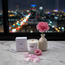On a marble table, the Tadpole & Piglet Damson & Rose Scented Candle with its box sits beside a vase with a pink carnation and scattered rose petals, all next to a window overlooking the city’s blurred night lights.