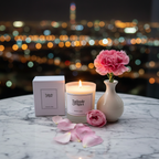 A Tadpole & Piglet Damson & Rose Scented Candle rests on a marble table beside its box, a white vase with a pink carnation, and rose petals, while city lights glow softly in the background to enhance its fruity-floral scent at night.