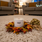 A Tadpole & Piglet Dark Amber & Ginger Lily Scented Candle rests on autumn leaves and acorns atop a light carpet, with dried flowers nearby. Cozy living room scene features gray armchairs and warm lighting in the background.