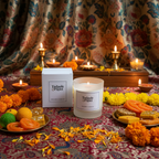 A lit Tadpole & Piglet Dark Amber & Ginger Lily Scented Candle sits beside its box, surrounded by marigolds, Indian sweets, and oil lamps on a patterned cloth with a floral curtain in the background.