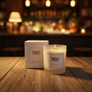 A lit Tadpole & Piglet Dark Honey & Tobacco Scented Candle with a wooden wick sits on a wooden table in a cozy bar, its box beside it. Warm lights and blurred shelves in the background create a relaxed, inviting atmosphere.