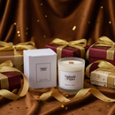 A Tadpole & Piglet Dark Honey & Tobacco Scented Candle (Seconds) and its box rest on brown fabric, surrounded by festively wrapped gifts with gold ribbons. The scene is both elegant and inviting.